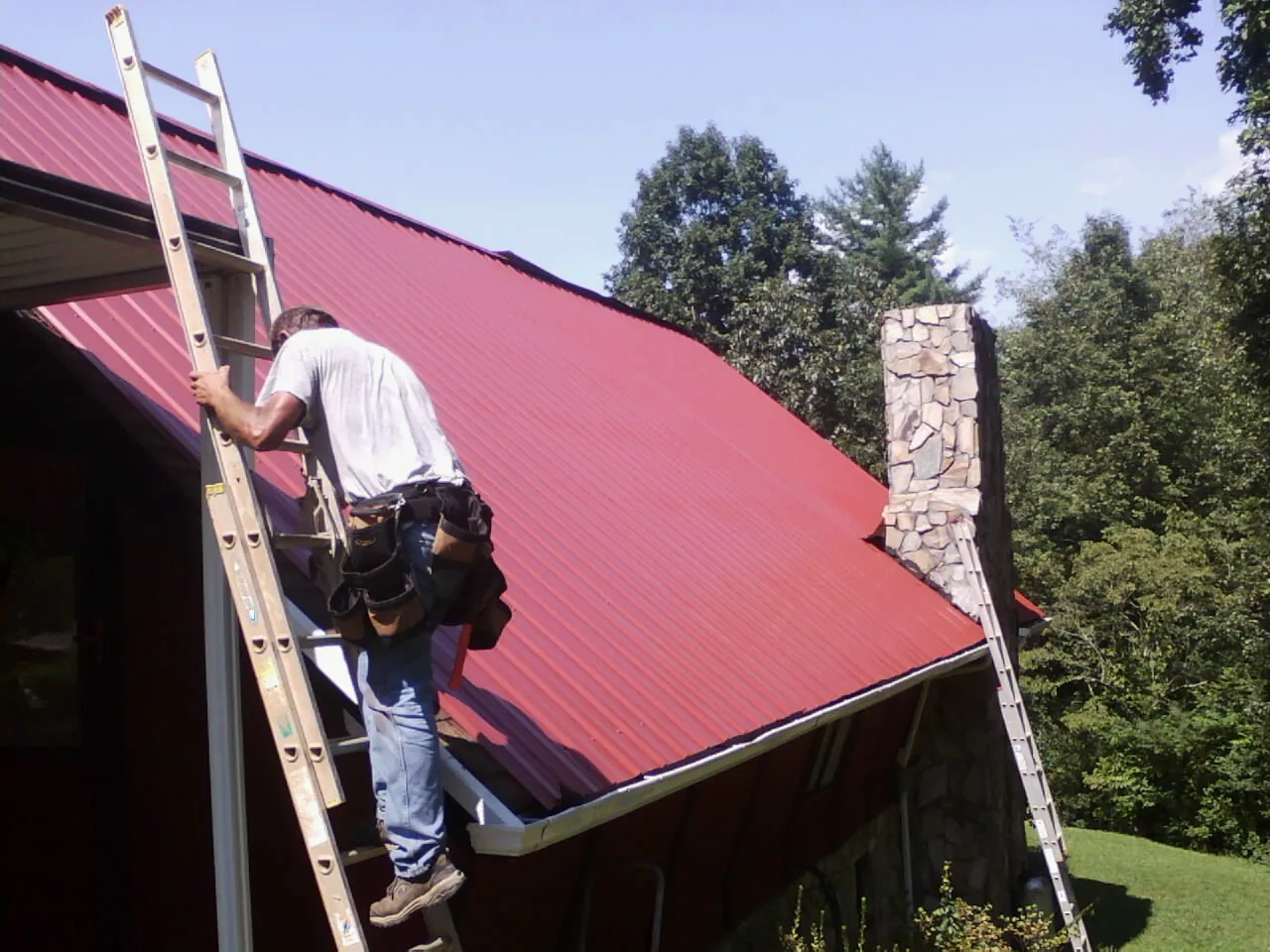 Roofer on a ladder beside a red metal roof and stone chimney — Soffit & Fascia Repair by Roofing Brothers in North Chelmsford